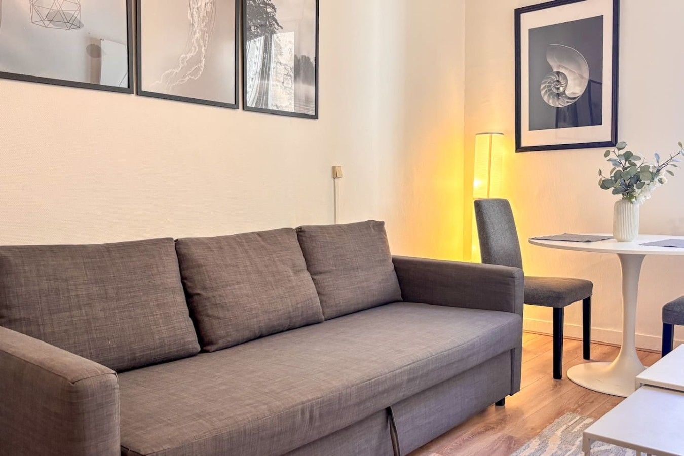 Un salon moderne avec un canapé gris, une petite table ronde surmontée d’un vase et des œuvres d’art encadrées accrochées au mur.