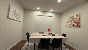 Comedor con una mesa preparada para cuatro personas, una ventana con persianas y dos cuadros colgados en la pared.