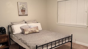 Un dormitorio con cama, mesita de noche, ventilador de pared, ventana con persianas y luz de techo.