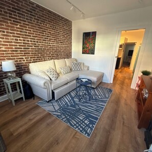 A living room with a sectional sofa, a small side table, a blue patterned rug, and a brick wall. - THE ARTFUL TIDE - Unit B/Beautiful 2 BR/1BA Mansion Suite (Atlantic City)