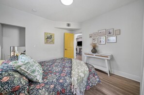 Ein Schlafzimmer mit Bett, Nachttisch, Spiegel, einer Tür und einer Wand mit gerahmten Bildern.