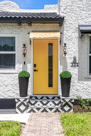 Un bâtiment avec une porte jaune, deux plantes en pot et un porche décoré de carreaux.