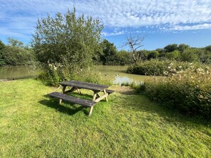 A picnic table in a grassy area with a pond and trees in the background. - Cottage Sleeps 4 Indoor Pool, Fishing Lake, Pets (Beaworthy)