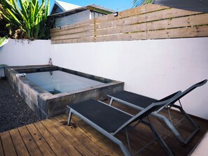 A hot tub with a wooden deck and lounge chairs, surrounded by a white wall and a wooden fence.