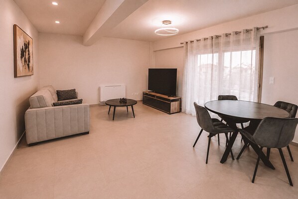 A modern living room with a sofa, a coffee table, a dining table with chairs, a flat-screen TV, and a window with curtains.
