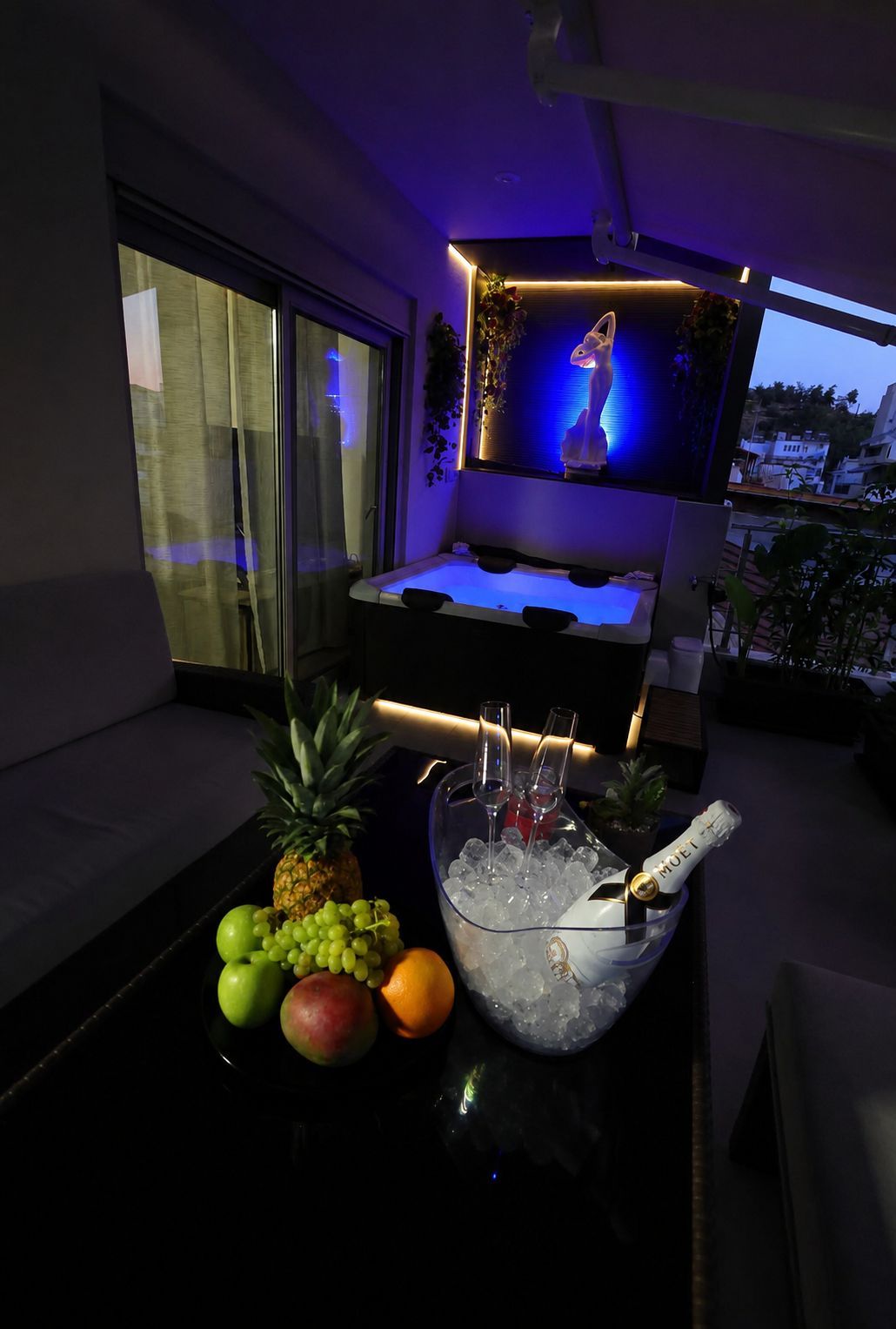 Ein Zimmer mit großem Fenster, einer Minibar mit einer Champagnerflasche und einer Obstschale mit Trauben und Ananas.
