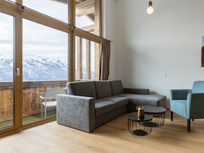 Ein modernes Wohnzimmer mit einem grauen Ecksofa, einem runden schwarzen Tisch mit einer Goldvase und einem blauen Sessel. Große Fenster bieten Blick auf schneebedeckte Berge.
