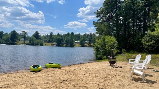 Eine Seenlandschaft mit Sandstrand, einem Ruderboot, einem Grill und Adirondack-Stühlen.