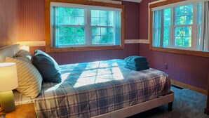 Ein Schlafzimmer mit einem Bett, einem Nachttisch, einer Lampe und zwei Fenstern mit Blick auf Grünflächen.