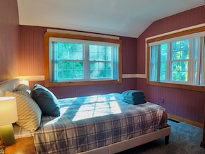 Une chambre à coucher comprenant un lit, une table de chevet, une lampe et deux fenêtres donnant sur un paysage verdoyant.