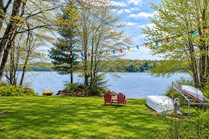 A lakeside setting with a grassy lawn, red Adirondack chairs, a canoe, and a boat docked near the water.