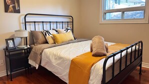 Cama bem arrumada com cabeceira, mesa de cabeceira com abajur e janela com vista para uma casa.