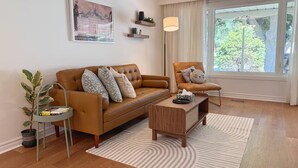 Uma sala de estar moderna com um sofá de couro marrom, uma mesa lateral redonda com uma planta, uma mesa de centro de madeira e uma janela grande com cortinas.