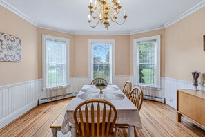 Ein Esszimmer mit langem Tisch, Stühlen, einer Chandelüster und großen Fenstern.
