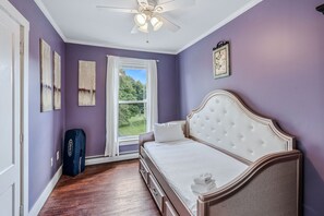 Ein Schlafzimmer mit einem weißen, gepolsterten Bett, einem Deckenventilator, einem Fenster mit Vorhängen und Blick ins Grüne.