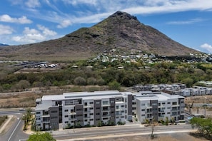 Un immeuble résidentiel moderne à plusieurs étages, doté d’un toit plat, entouré d’une route et d’un paysage vallonné à la végétation clairsemée.