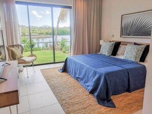 A bedroom with a bed, a desk, and a chair. There is a large window with a view of greenery and water. - Luxury Waterfront Apartment with Pools (Grande Riviere Noire)