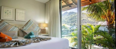 A hotel room with a bed, a nightstand, a lamp, and a large window offering a view of a mountainous landscape and palm trees.