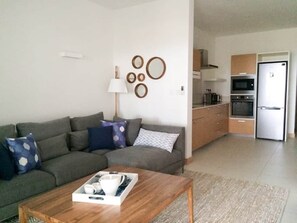 Ein Wohnzimmer mit einer Couch, einem Couchtisch und Wanddekoration. Im Hintergrund ist eine Küche mit Geräten zu sehen.