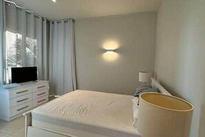 A hotel room with a bed, a bedside lamp, a television on a dresser, and a window with curtains. - Waterfront Apartment in Marina (Rivière Noire District)