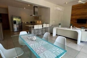 Uma cozinha moderna com eletrodomésticos de aço inoxidável, uma sala de jantar com mesa de vidro e uma sala de estar com televisão.