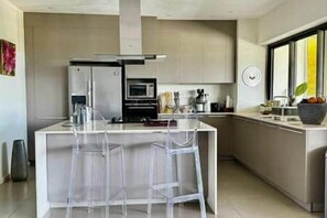 A modern kitchen with a central island, bar stools, stainless steel appliances, and a large window providing natural light. - Penthouse with Panoramic Sea Views (Tamarin)