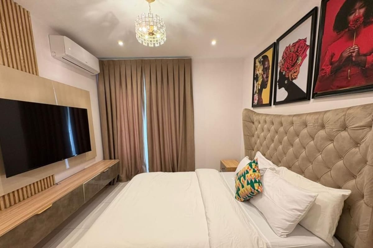 Quarto de hotel com uma televisão de tela plana grande, cabeceira estofada, cama com roupa de cama branca e ar-condicionado fixado na parede.