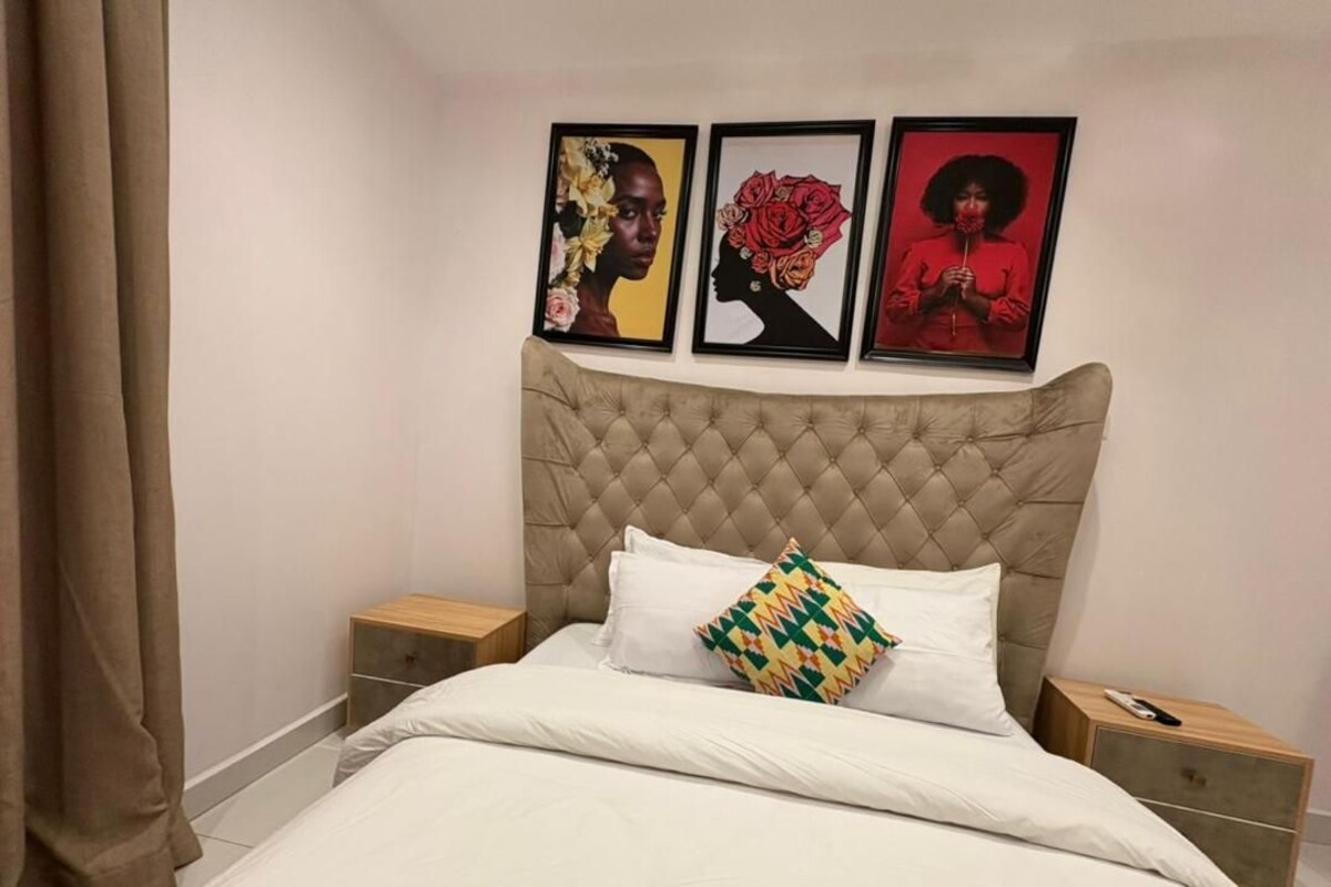 Quarto com cabeceira estofada, roupa de cama branca, almofada colorida e três obras de arte emolduradas na parede.