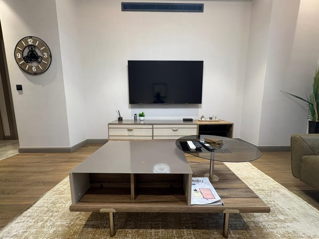 A modern living room with a flat-screen TV mounted on the wall, a wooden coffee table with a magazine, and a glass side table with a remote control.