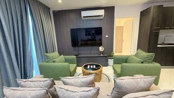 A modern living room with a flat-screen TV, green sofas, a coffee table, and a dining area with dark wood cabinets.