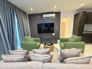 Uma sala de estar moderna com televisão de tela plana, sofás verdes, uma mesa de centro e um espaço de jantar com armários de madeira escura.