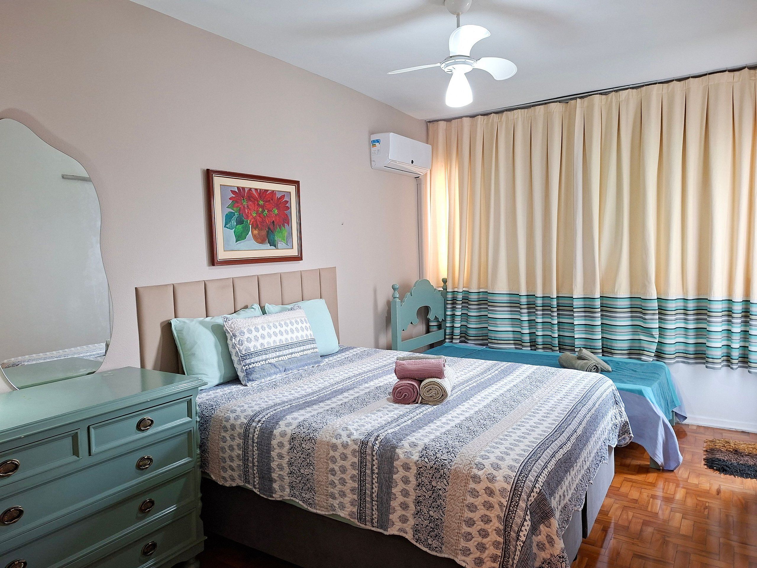 Un dormitorio con cama, cómoda, ventilador de techo, ventana con cortinas y aire acondicionado instalado en la pared.