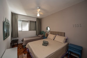Un dormitorio con dos camas, un ventilador de techo, una ventana con cortinas, una mesita de noche y un pequeño aparador.
