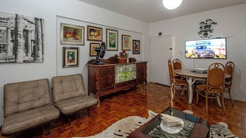 Una sala de estar con un conjunto de sofás, una mesa de comedor con sillas, un televisor montado en la pared y diversas obras de arte enmarcadas.