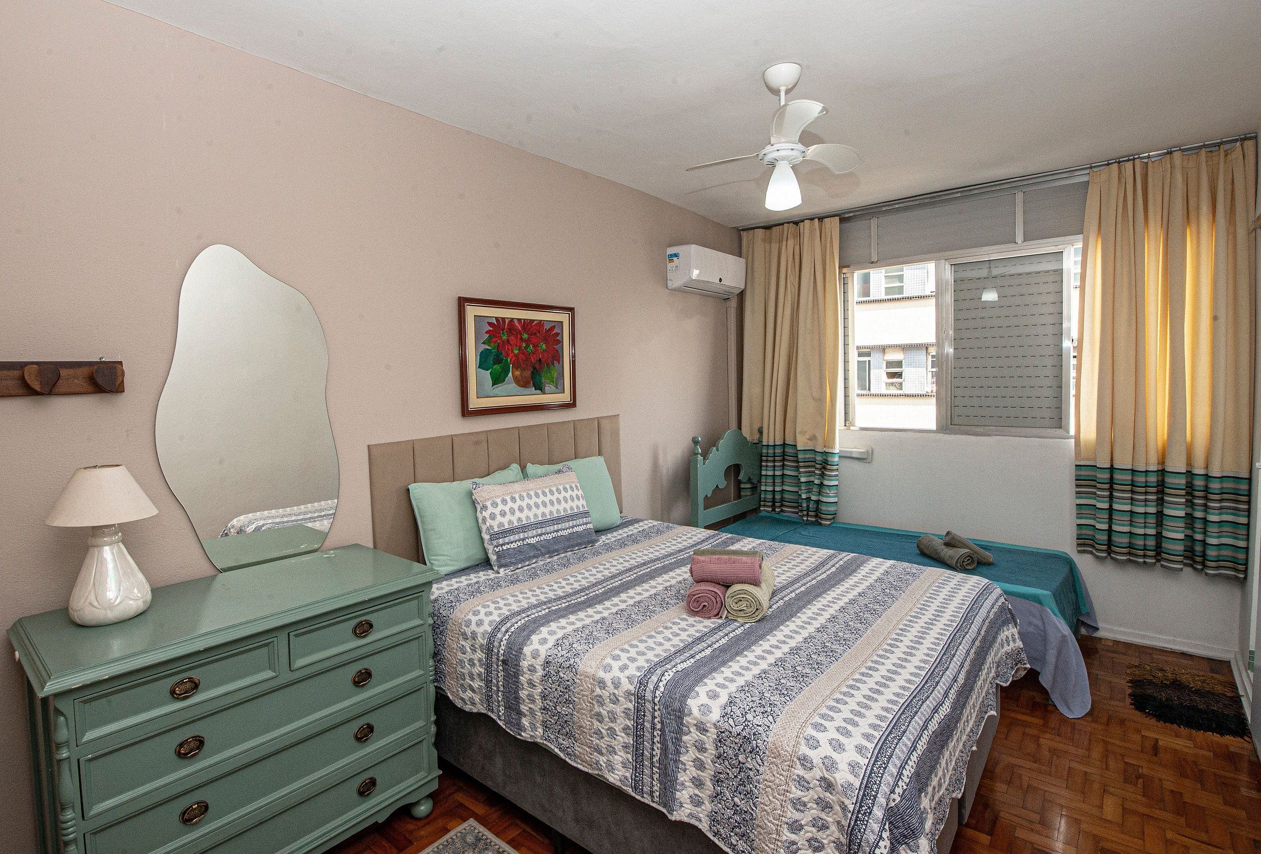 Un dormitorio con cama, cómoda, ventilador de techo, ventana con cortinas y un cuadro en la pared.