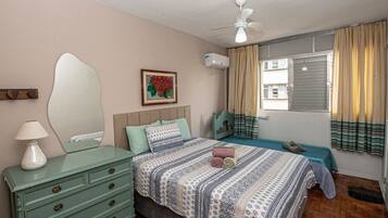 Un dormitorio con cama, cómoda, ventilador de techo, ventana con cortinas y un cuadro en la pared.