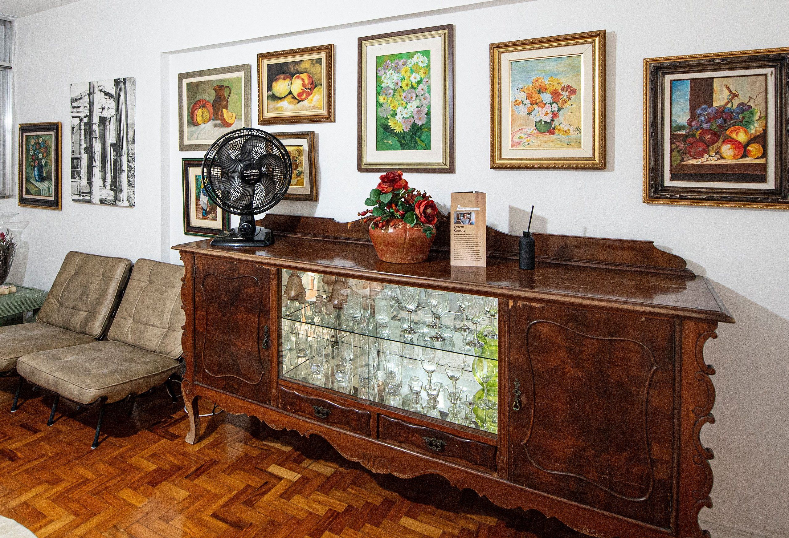 A room with a wooden cabinet displaying glassware, a fan, and a vase with flowers. There are framed artworks on the wall.