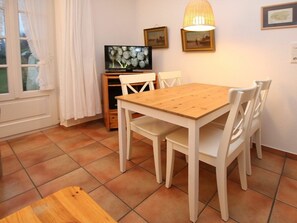 A dining area with a wooden table and white chairs, a television on a stand, and a lamp on the wall. - 1, Dorpstraat 9 (Wyk auf Foehr)