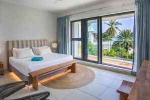 Ein Hotelzimmer mit einem großen Bett, einer Holzbank, einem runden Teppich und Blick auf Palmen durch große Fenster.