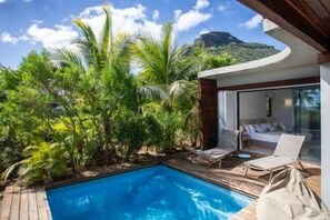A poolside area with a wooden deck, lounge chairs, and a glass-enclosed room with a view of a mountain. - Beachfront Villa with Private Pool & Garden (Black River)