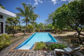 A rectangular swimming pool with a tiled edge, surrounded by a stone patio. There are palm trees and other greenery in the background. - The Villa - Le Morne (Le Morne)