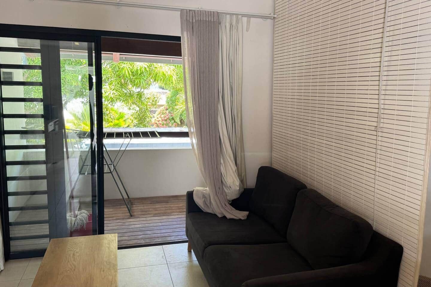 A room with a sliding glass door leading to a balcony, a dark-colored sofa, and a window with blinds.