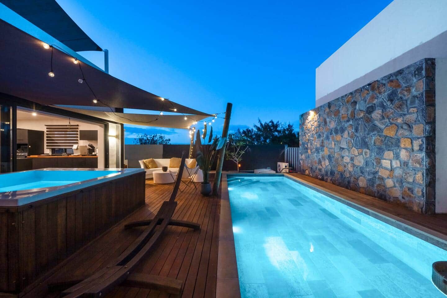 A modern outdoor pool area with a hot tub, wooden deck, and stone wall.