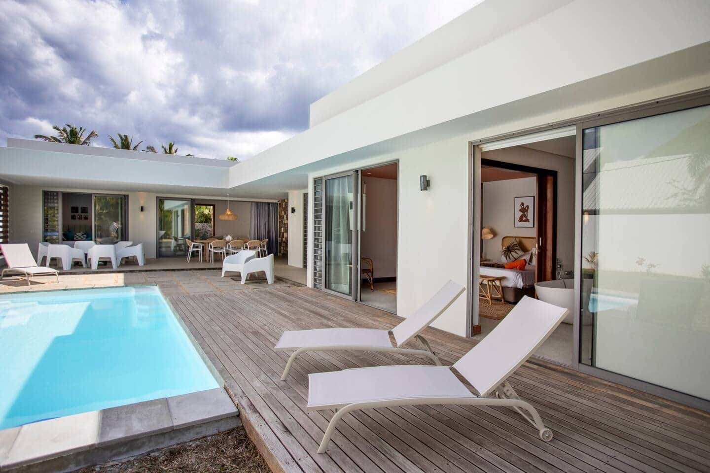 A modern outdoor pool area with lounge chairs, a dining area, and a bedroom with a bathtub.