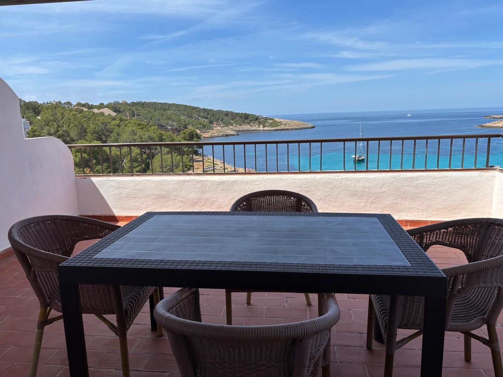 A terrace with a table and chairs, overlooking a coastal view with a clear sky.