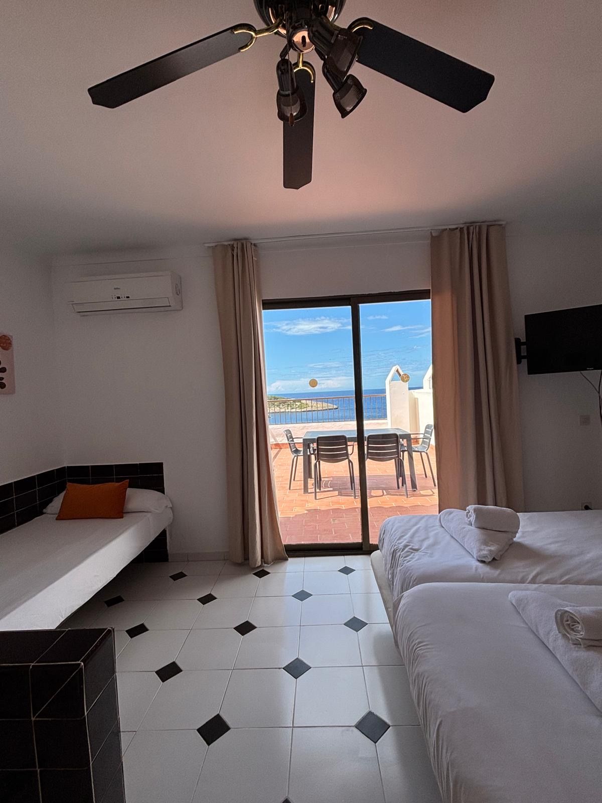 A hotel room with two beds, a ceiling fan, an air conditioning unit, a TV, and a view of the sea through a sliding glass door.