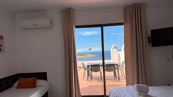 A hotel room with two beds, a ceiling fan, an air conditioning unit, a TV, and a view of the sea through a sliding glass door.