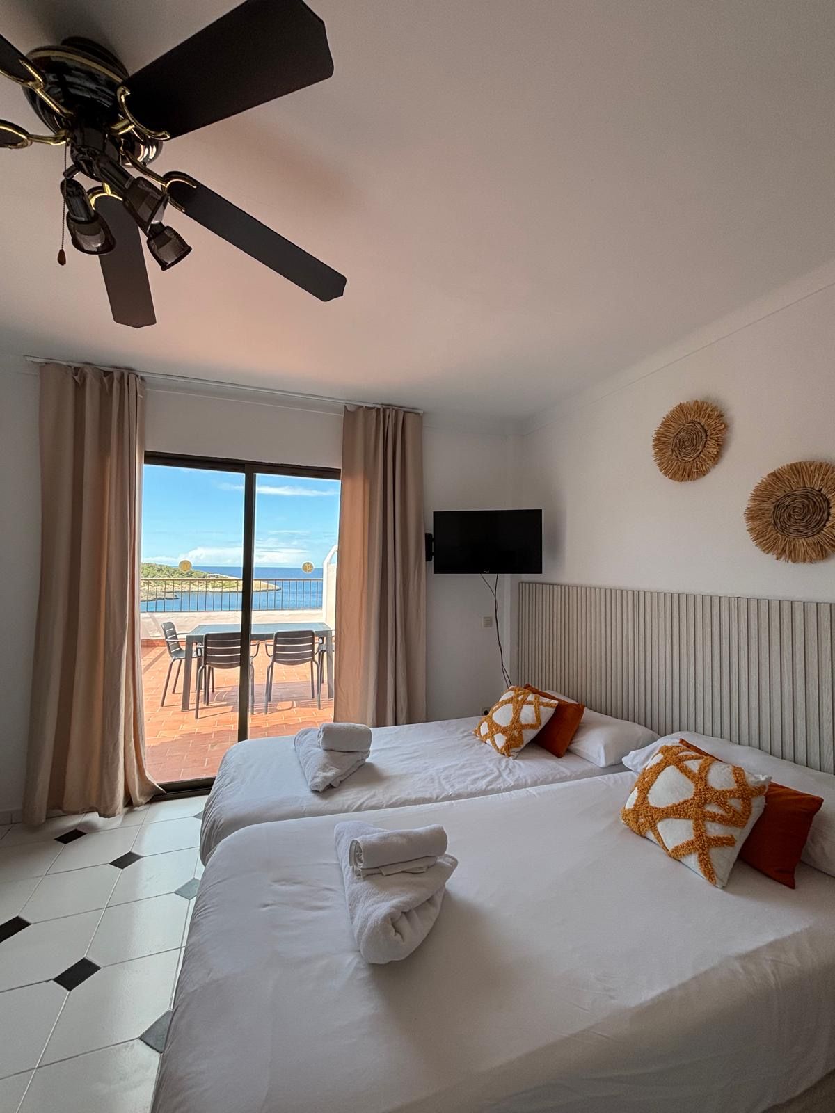 A hotel room with two beds, a ceiling fan, a TV, and a balcony with a view of the sea.