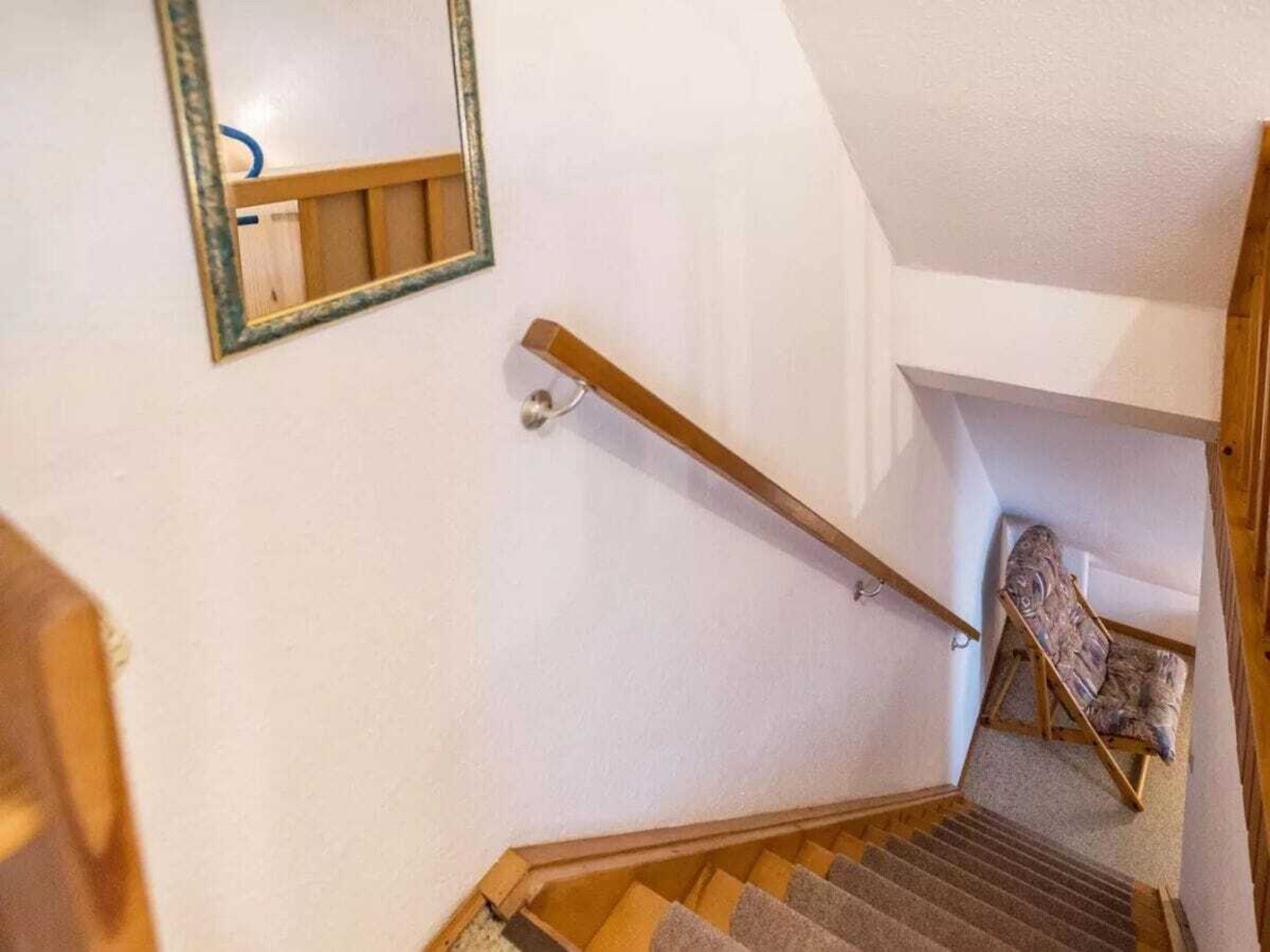 Uma escada com corrimão de madeira, uma cadeira na base e um espelho na parede.