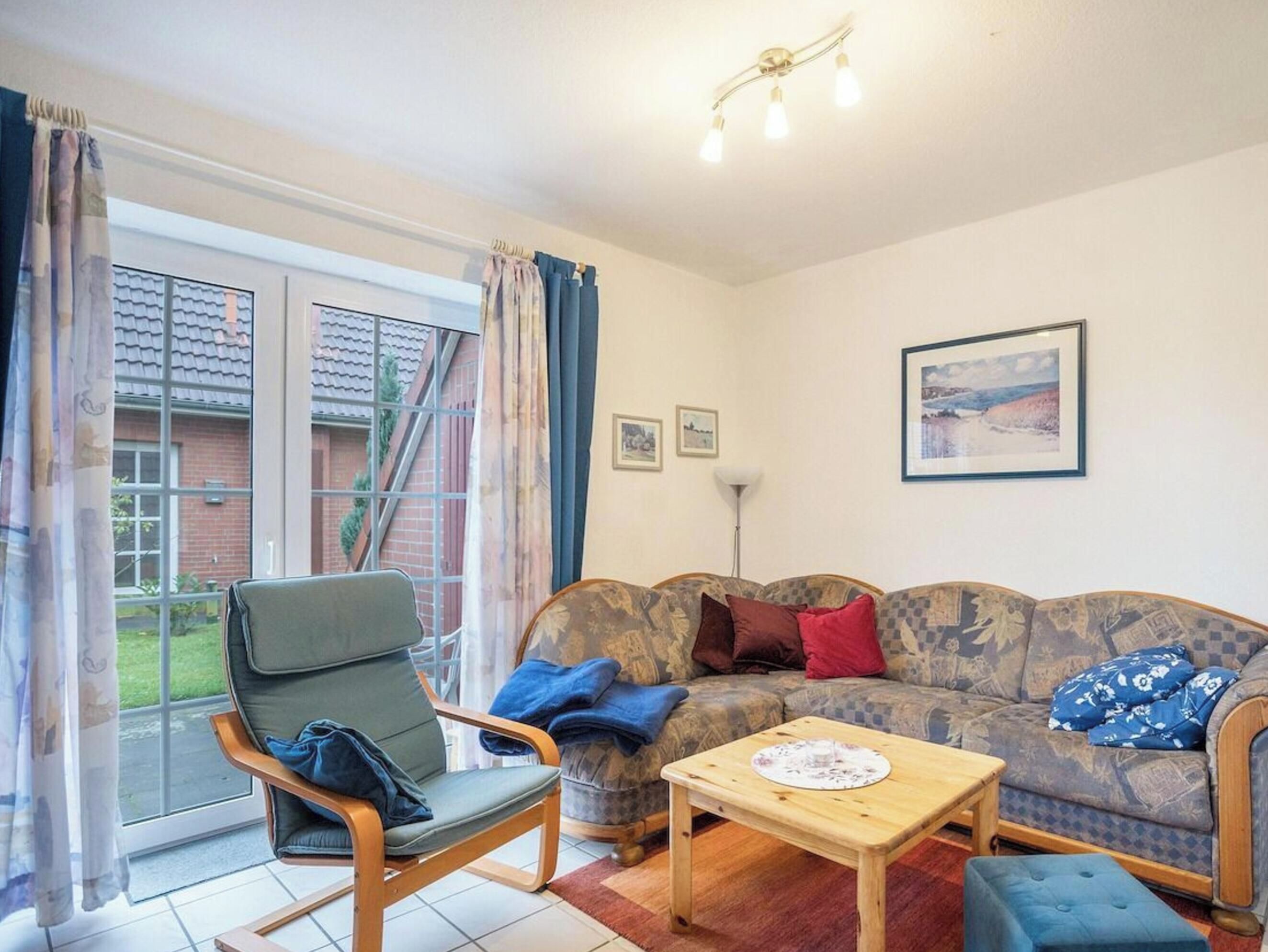 Een woonkamer met een bank, fauteuil en een kleine tafel. Er is een raam met gordijnen, een lamp en een ingelijst schilderij aan de muur.